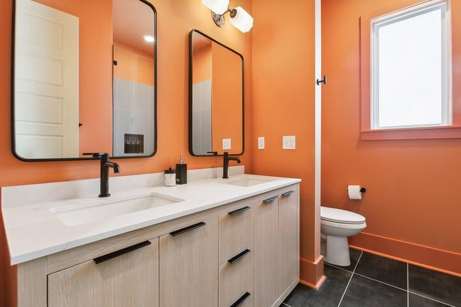 Vibrant orange bathroom featuring a white quartz double vanity with black faucets and rectangular mirrors, light wood cabinets, black floor tiles, and a toilet beneath a frosted window.