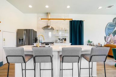 Bright modern kitchen with a white island and four gray bar stools, brass gooseneck faucet, stainless steel fridge and range, marble backsplash with open shelves, gold linear pendant light and deep teal curtain