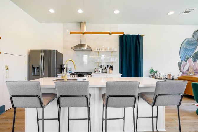 Bright modern kitchen with a white island and four gray bar stools, brass gooseneck faucet, stainless steel fridge and range, marble backsplash with open shelves, gold linear pendant light and deep teal curtain