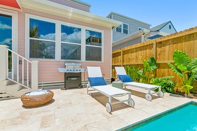Sunny residential poolside patio with two white lounge chairs and blue pillows, a stainless-steel grill by a pastel pink house, tropical plants, wooden privacy fence and turquoise pool water.