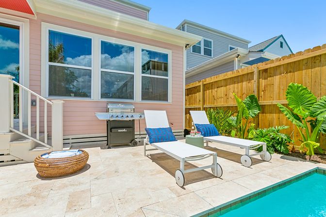 Sunny residential poolside patio with two white lounge chairs and blue pillows, a stainless-steel grill by a pastel pink house, tropical plants, wooden privacy fence and turquoise pool water.