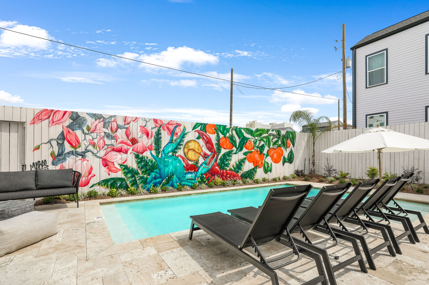 Sunny residential backyard swimming pool with a row of black lounge chairs and white umbrella on a stone deck, set against a vibrant wooden-fence mural of oversized pink flowers, green foliage, bright oranges and a blue crab under a clear blue sky.