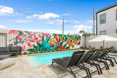 Sunny residential backyard swimming pool with a row of black lounge chairs and white umbrella on a stone deck, set against a vibrant wooden-fence mural of oversized pink flowers, green foliage, bright oranges and a blue crab under a clear blue sky.