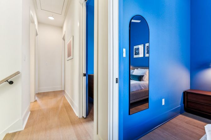 Modern home interior with a bright white hallway opening to a vivid blue bedroom, an arched full-length mirror reflecting a bed with pillows, a wood nightstand, and light hardwood floors.