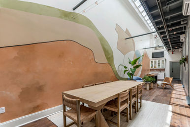 Sunlit contemporary loft dining area with a long wooden table and woven chairs, abstract pastel wall mural, skylight and exposed industrial ceiling with potted plants