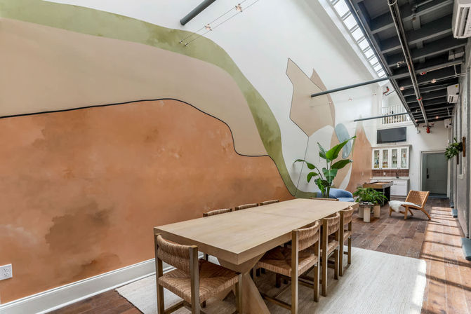 Sunlit contemporary loft dining area with a long wooden table and woven chairs, abstract pastel wall mural, skylight and exposed industrial ceiling with potted plants