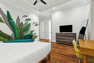 Bright modern bedroom with oversized tropical leaf wall mural, crisp white bed with teal throw, hardwood floors, dresser with TV, leaning mirror and wooden desk with green chair.