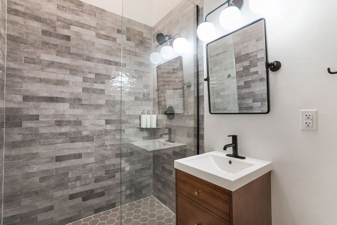 Modern bathroom with glass walk-in shower, gray subway-style wall tile and hexagon floor tile, wood vanity with white sink and black faucet, black-framed mirror and globe vanity lights.