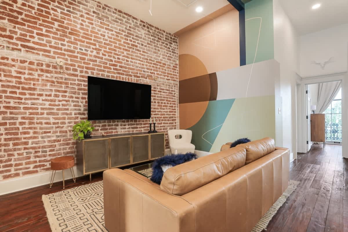 Sunlit urban loft living room with exposed red brick wall, wall-mounted TV, tan leather sofa, geometric pastel mural, hardwood floors and patterned rug.