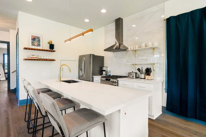 Bright modern open-plan kitchen with large white quartz island, gold faucet, gray upholstered bar stools, stainless steel refrigerator and range, marble-look backsplash, open shelves with glassware, brass accents, pendant light, and hardwood floors