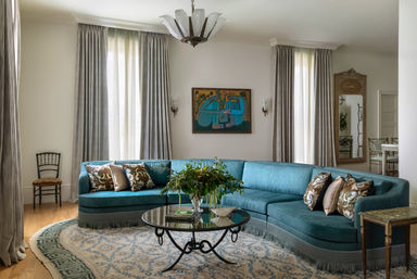 Bright elegant living room with a teal curved sectional sofa adorned with patterned throw pillows, round glass coffee table topped with a leafy centerpiece, tall draped windows, decorative wall art, chandelier and a patterned area rug — stylish home interior.