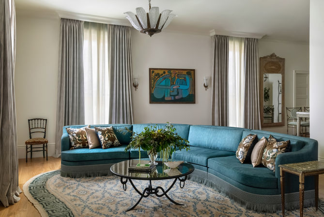 Bright elegant living room with a teal curved sectional sofa adorned with patterned throw pillows, round glass coffee table topped with a leafy centerpiece, tall draped windows, decorative wall art, chandelier and a patterned area rug — stylish home interior.