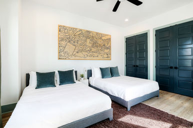 Bright modern bedroom with two beds in white linens, teal throw pillows, whimsical wall art above the headboards, dark teal paneled closet doors, ceiling fan, light wood floors and a plush brown rug