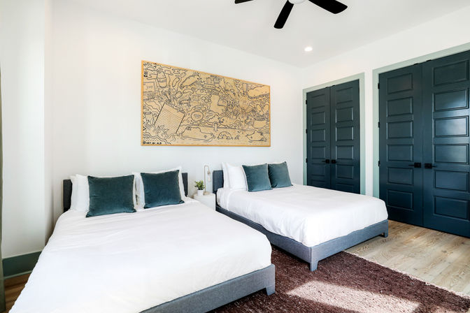 Bright modern bedroom with two beds in white linens, teal throw pillows, whimsical wall art above the headboards, dark teal paneled closet doors, ceiling fan, light wood floors and a plush brown rug