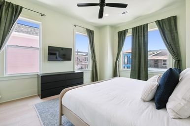 Bright modern bedroom with white bed and navy pillows, three large windows with green curtains overlooking colorful urban houses, wall-mounted TV, black dresser, ceiling fan and light hardwood floors.
