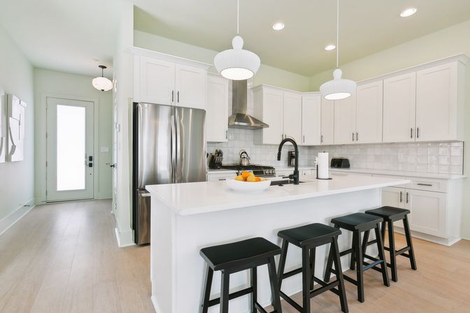 Bright modern white open-concept kitchen with quartz island, four black bar stools, stainless-steel appliances, pendant lights and light hardwood floors.