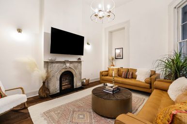 Cozy high‑ceiling living room in an urban townhouse with a marble fireplace and wall‑mounted TV, two caramel leather sofas, round wood coffee table on a patterned rug, pampas grass, potted plant, and modern chandelier.
