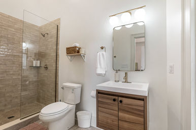 Bright modern residential bathroom with frameless glass walk-in shower, beige subway tile, white toilet, wood vanity with white sink and round-corner mirror