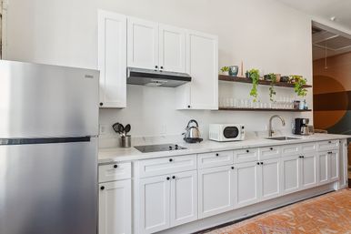 Bright modern white open-concept kitchen with stainless steel refrigerator, marble countertop, electric cooktop, microwave and coffee maker, floating wood shelves with glassware and trailing plants.
