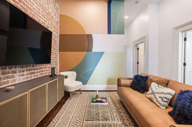 Contemporary living room with exposed brick TV wall, large colorful geometric mural, tan leather sofa with textured and blue fur pillows, patterned area rug, glass coffee table, and a white accent chair.