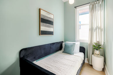 Bright, cozy small bedroom with a navy velvet daybed, white quilted bedding and a teal pillow against seafoam green walls, abstract black-and-white art, sunlit window with sheer curtains and a potted plant on a ribbed side table.
