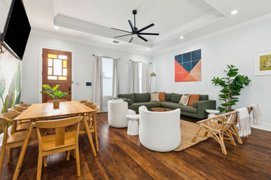 Bright modern open-plan living and dining room with hardwood floors, green sectional sofa, wooden dining table, rattan chairs, large ceiling fan, indoor plants, and geometric wall art.