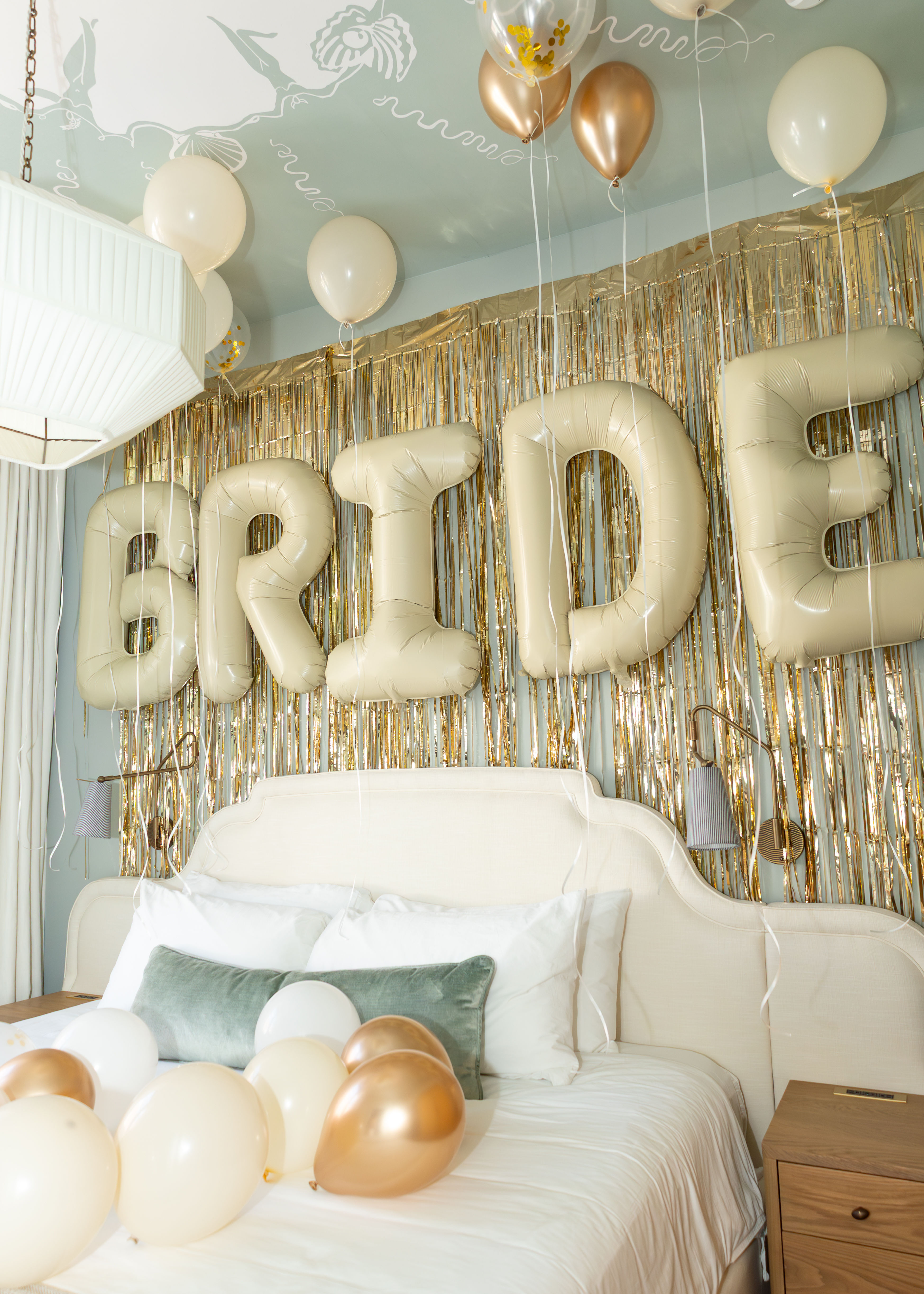 Bridal suite with giant gold 'BRIDE' letter balloons, metallic gold fringe backdrop and cream and rose-gold balloons over a white bed