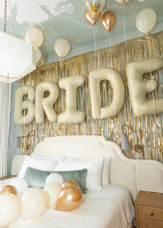 Bridal suite with giant gold 'BRIDE' letter balloons, metallic gold fringe backdrop and cream and rose-gold balloons over a white bed