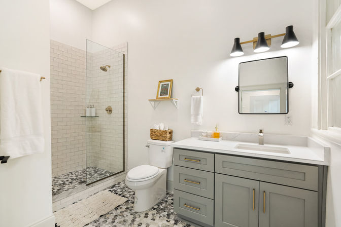Bright modern residential bathroom with glass walk-in shower and white subway tile, gray-and-white mosaic floor, gray vanity with marble countertop and brass hardware, wall mirror, triple vanity lights, towel and wicker storage basket.
