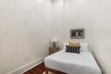 Cozy minimalist bedroom nook with white walls and dark hardwood floor, single bed dressed in white linens with yellow and striped accent pillows, small wooden nightstand with green lamp, and framed black-and-white photograph on the wall.