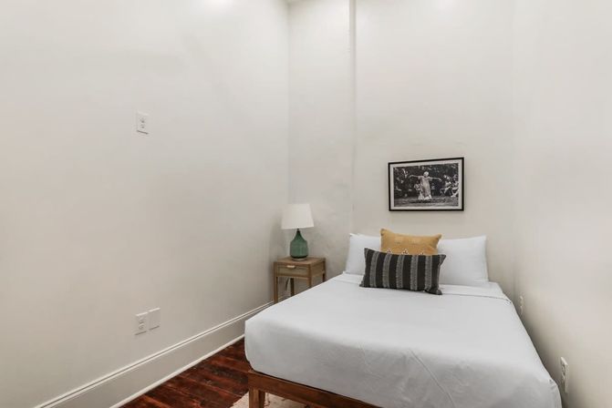 Cozy minimalist bedroom nook with white walls and dark hardwood floor, single bed dressed in white linens with yellow and striped accent pillows, small wooden nightstand with green lamp, and framed black-and-white photograph on the wall.