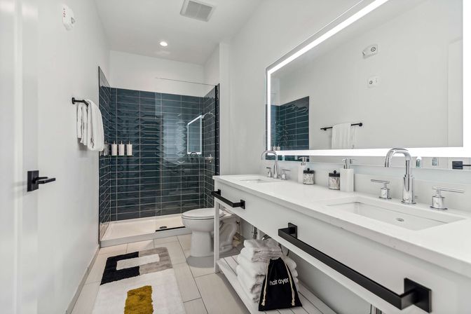 Bright modern bathroom interior with white double vanity and LED-lit mirror, chrome faucets, open shelf with folded towels and hair-dryer bag, glass walk-in shower with dark blue subway tile and built-in niches, toilet and patterned bath rug.