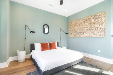 Sunlit modern bedroom with mint-green walls, low gray platform bed dressed in white linens and rust-orange pillows, black swing-arm wall lamps, oval mirror, large framed doodle artwork, and hardwood floors.