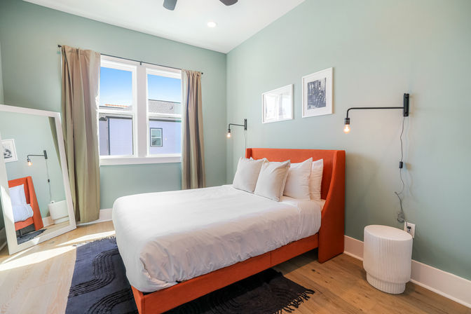 Sunlit townhouse bedroom with orange upholstered bed, white linens, seafoam-green walls, Edison wall lamps, full-length mirror and hardwood floors