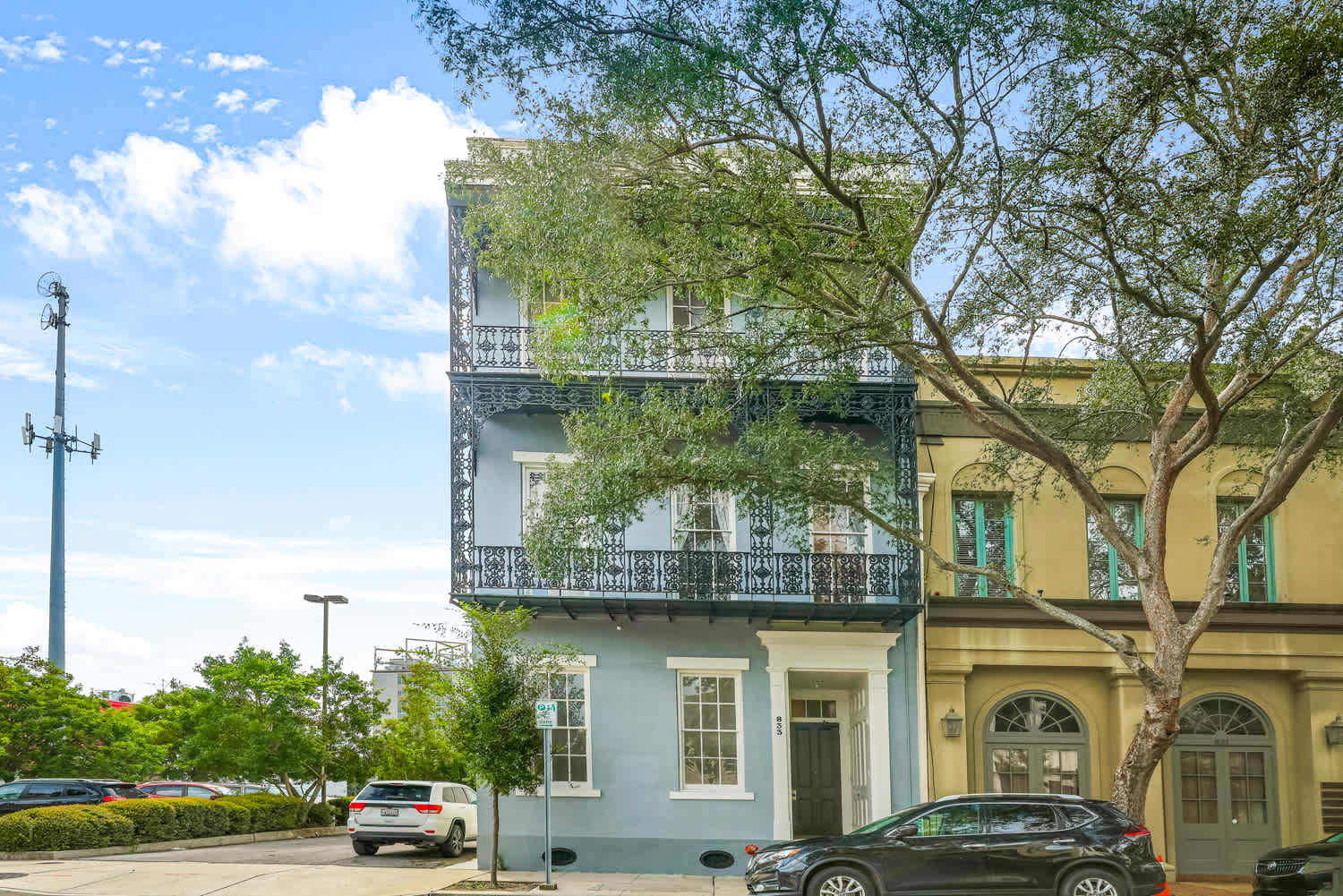 Pastel blue three-story rowhouse with ornate wrought-iron balconies on a tree-lined street, neighboring tan building and parked cars under a bright blue sky.