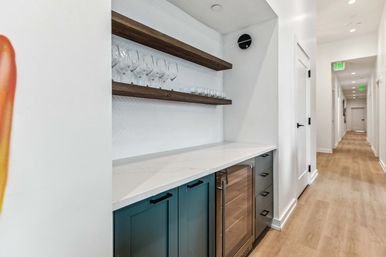 Sleek hallway beverage nook with white quartz countertop, teal lower cabinets and glass-front wine cooler, two wooden floating shelves holding wine and cocktail glasses, white herringbone backsplash and light wood flooring leading down a bright corridor.