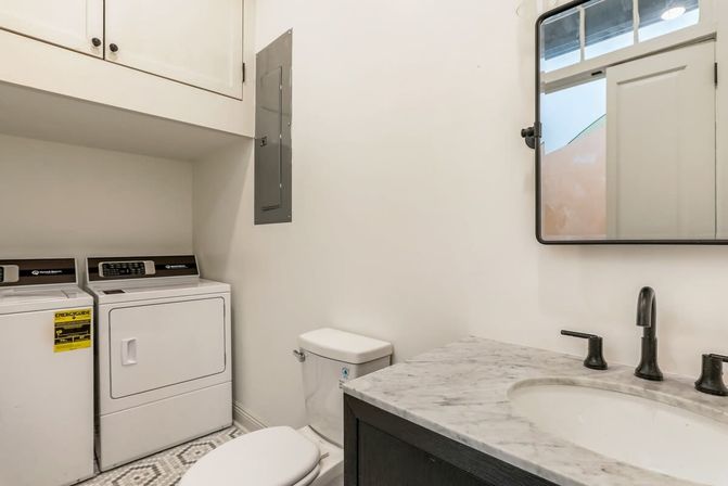 Compact laundry-bath combo with side-by-side washer and dryer, white toilet, marble-top vanity with black faucet and mirror, upper cabinets and patterned tile floor.