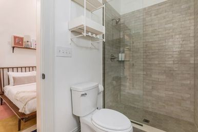 Bright apartment bathroom with white toilet, wall-mounted shelf, and glass walk-in shower with beige subway-style tile; bedroom visible through open door