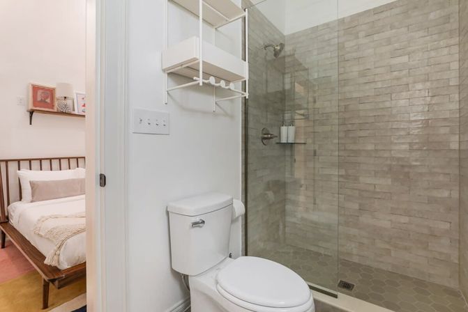 Bright apartment bathroom with white toilet, wall-mounted shelf, and glass walk-in shower with beige subway-style tile; bedroom visible through open door