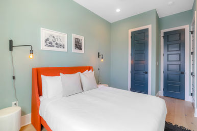 Bright modern bedroom with soft teal walls, orange upholstered headboard providing a pop of color, crisp white bedding and pillows, black wall-mounted Edison-bulb sconces, framed black-and-white photos, and two dark blue paneled doors.