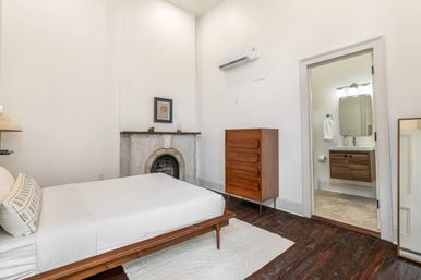 Bright, minimalist bedroom interior with white walls, low mid-century wooden bed, textured rug, vintage marble fireplace, tall wooden dresser, dark hardwood floors, wall-mounted AC and open doorway to a modern en-suite bathroom.