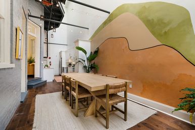 Bright urban loft dining area with a long light-wood table and woven chairs, open kitchen, abstract terracotta-and-green mural, high industrial ceiling, pendant lights, hardwood floors and indoor plants.
