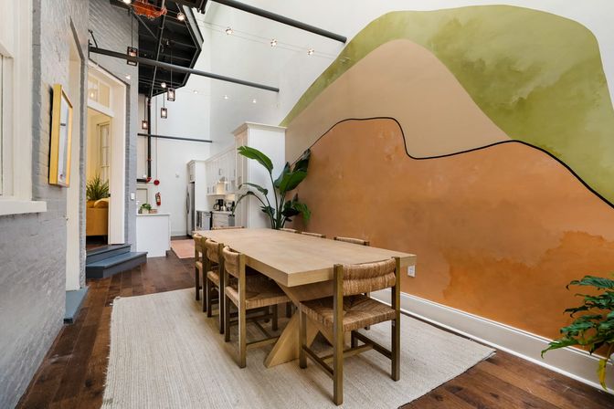 Stylish urban loft dining area with long wooden table and woven chairs by an abstract terracotta-and-green mural, open-concept kitchen, high ceilings and hanging pendant lights