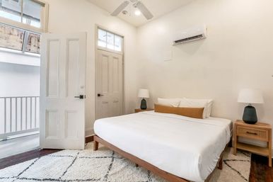 Bright minimalist apartment bedroom with white bedding on a wooden platform bed, matching bedside tables and lamps, ceiling fan, wall-mounted air conditioner and small balcony visible through an open door.