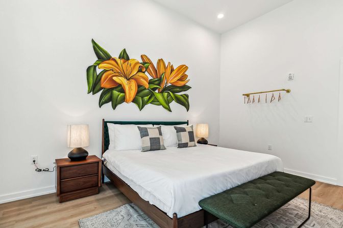 Bright modern bedroom with white walls, large orange lily mural above a mid-century wooden bed, crisp white bedding with checkered throw pillows, matching wooden nightstands and lamps, green upholstered bench and light wood floors
