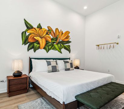Bright modern bedroom with white walls, large orange lily mural above a mid-century wooden bed, crisp white bedding with checkered throw pillows, matching wooden nightstands and lamps, green upholstered bench and light wood floors