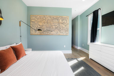 Sunlit modern bedroom with seafoam-green walls, white bed and rust-orange pillows, playful hand-drawn mural above the bed, black swing wall lamp, white dresser and hardwood floors.