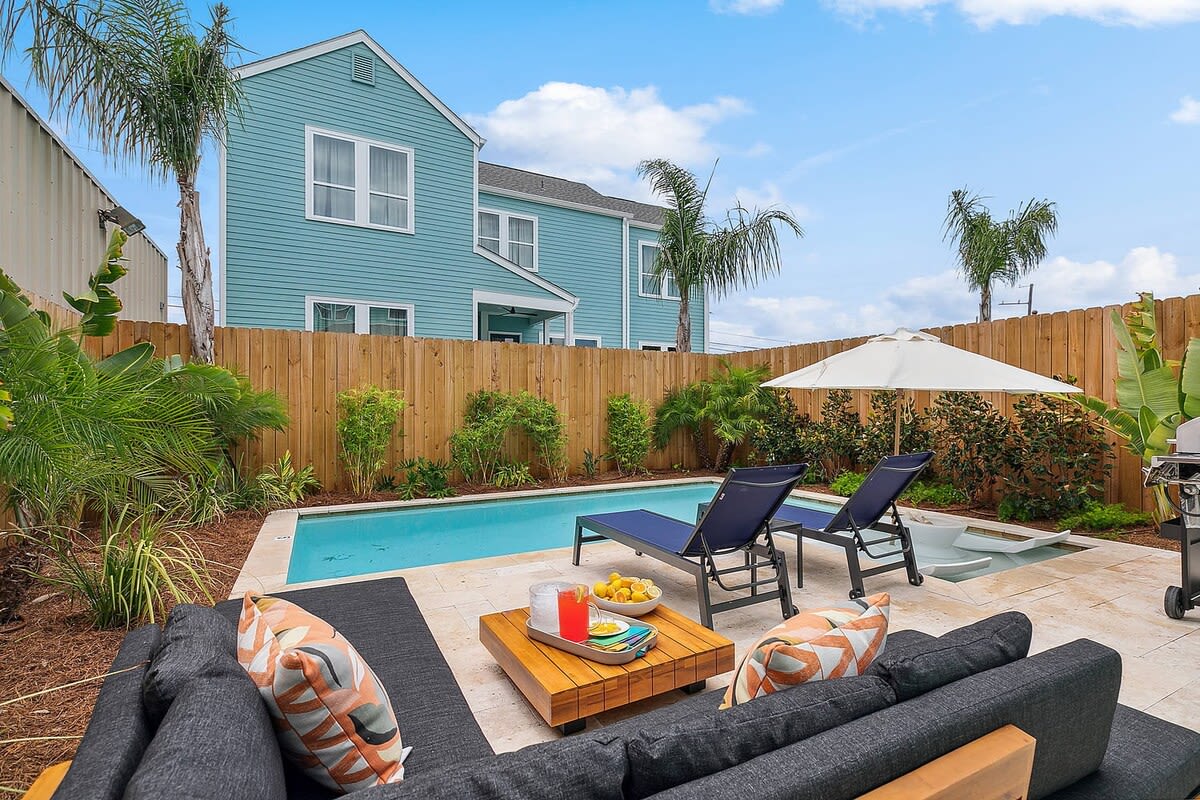 Backyard pool patio with a turquoise two-story house, wooden fence, palm trees, two navy lounge chairs under an umbrella, an outdoor sofa with colorful pillows and a small table with drinks and fruit.