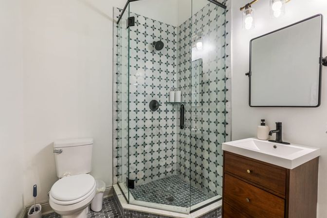Bright modern residential bathroom with glass-enclosed corner shower featuring patterned tiles, dark mosaic shower floor, wood vanity with white sink and black faucet, and a white toilet.