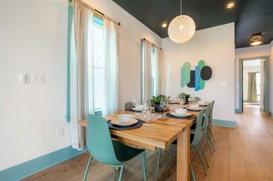 Sunlit modern dining room with a wooden table set for six, teal chairs and trim, round pendant light, abstract wall art and hardwood floors